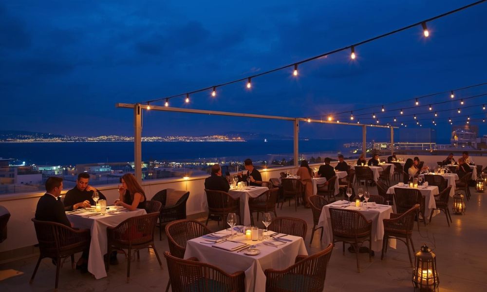 Breathtaking rooftop dining experience in tangier during blue hour showing elegant candlelit tables on a modern terrace with panoramic views across the strait of gibraltar to the spanish coast, twinkling medina lights below, string lights and traditional moroccan lanterns creating ambient lighting, sophisticated diners enjoying fusion cuisine, capturing the magical atmosphere where tangier's geographic position as a cultural crossroads meets contemporary culinary innovation.
