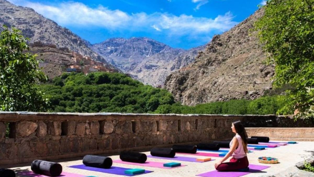A woman meditating on a sun-lit Atlas-facing terrace in Marrakech, yoga mats and cushions arranged around her, green valley and mountains in the background, reflecting calm, realism, and mindful slow travel.
