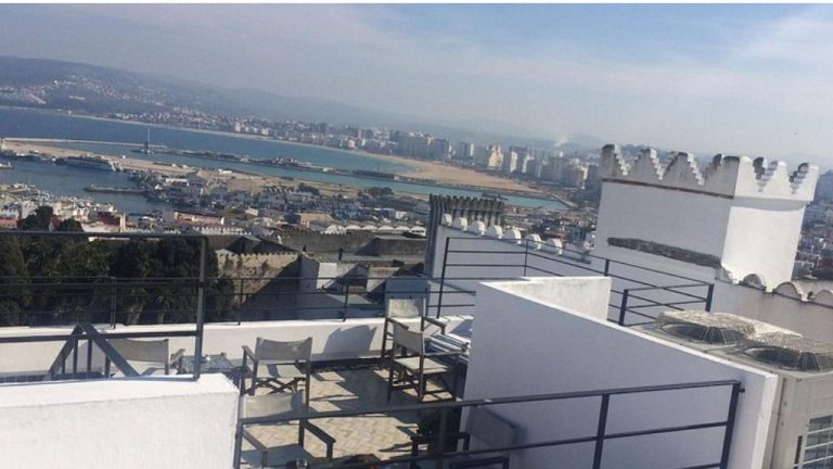 Modern minimalist rooftop terrace in tangier's medina featuring contemporary lounge chairs on white platforms overlooking breathtaking panoramic views of the strait of gibraltar, the beach, port area, and distant spanish coastline across the water. Traditional white moroccan architecture with crenellated walls frames the sleek outdoor furniture, capturing the blend of historic and modern design that characterizes tangier's upscale dining scene.