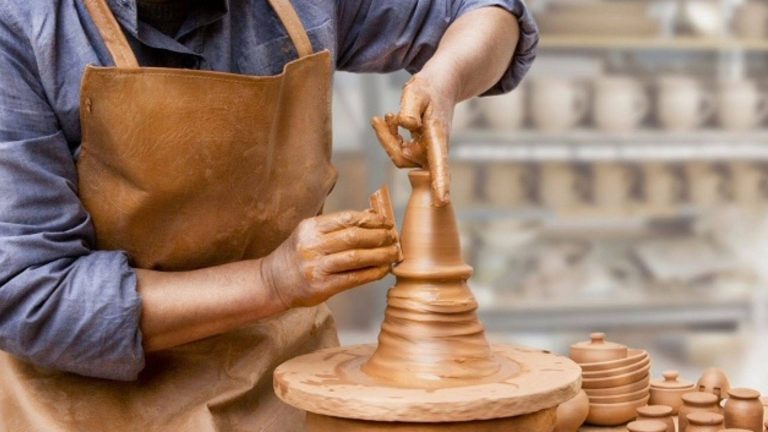 A Moroccan potter in Fes, wearing a leather apron, shapes wet red clay on a traditional foot-powered wheel his hands covered in earth, fingers guiding the rising vessel with quiet precision. In the background, stacked unfired tagines and bowls await the kiln, embodying a living tradition where silence is not absence, but presence shaped by hand, time, and memory.