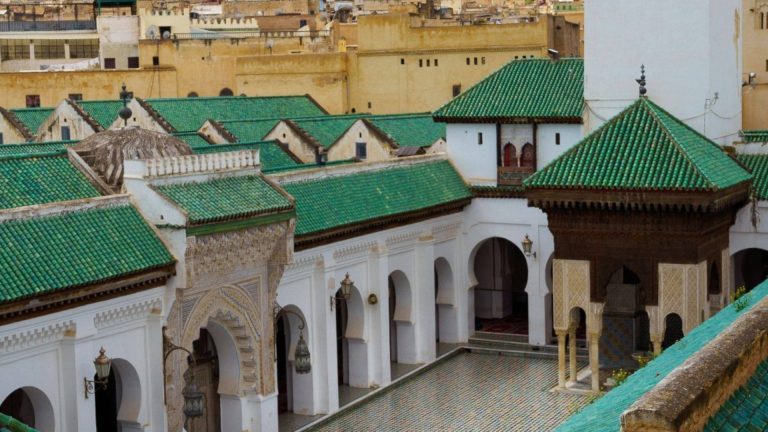 An aerial view of a historic riad courtyard in Fes, featuring vibrant green zellige tiled roofs, white plaster walls, arched colonnades, and a central fountain no people, no modern intrusions, just the quiet harmony of light, water, and geometry that defines Fes’ living tradition of architectural stillness.