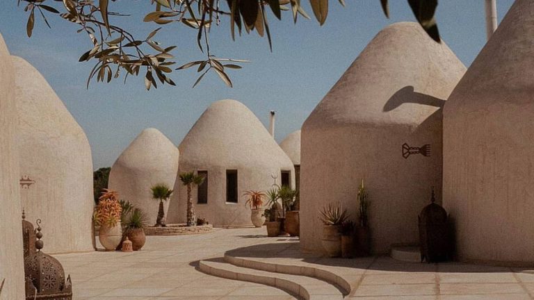 White dome-shaped riad buildings in Essaouira under a clear blue sky, surrounded by potted plants and olive branches embodying the quiet, earthy, and organic wellness culture of Morocco’s coastal retreat.
