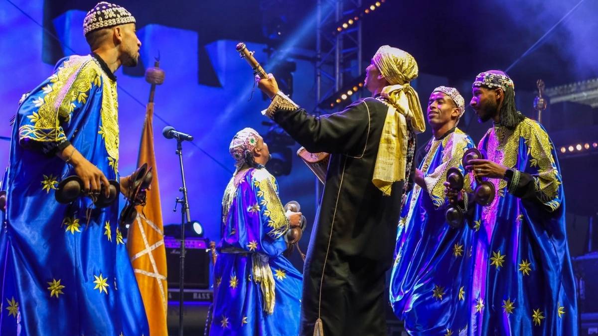 Four Gnaoua musicians in vibrant blue and gold djellabas perform on stage under dramatic blue lighting, holding qraqeb and guembri during the annual Essaouira World Music Festival a fusion of sacred ritual and global celebration.