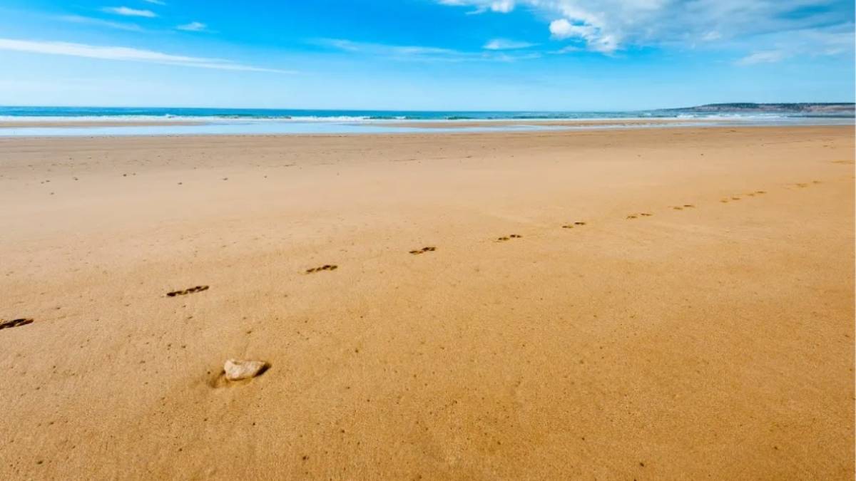 A serene, empty beach in Essaouira under a clear blue sky, with footprints trailing across golden sand toward the Atlantic waves symbolizing mindful walking, ocean-based healing, and the quiet rhythm of daily life by the sea.