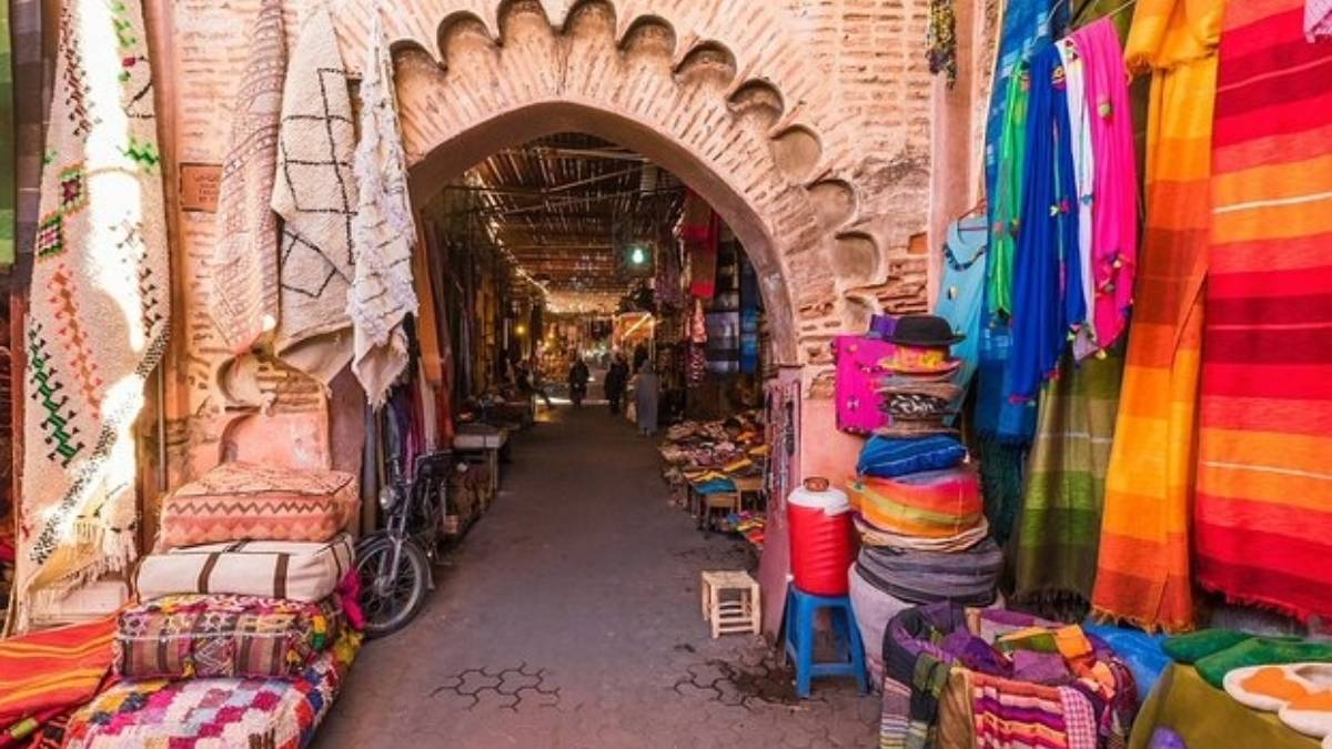 A vibrant alley in Essaouira’s medina framed by a traditional scalloped arch, lined with handwoven rugs, colorful textiles, and artisan stalls under warm afternoon light symbolizing mindful walking, cultural presence, and the slow rhythm of daily life in Morocco