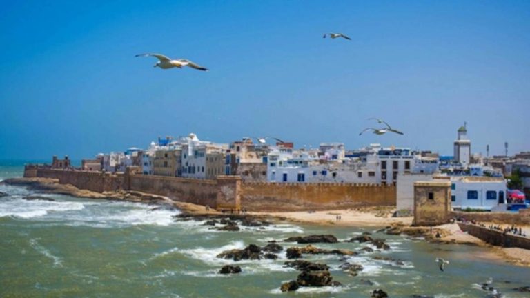 Historic coastal city of Essaouira with ancient stone walls and whitewashed buildings overlooking the Atlantic Ocean, seagulls flying above the waves, reflecting seasonal wellness, wind, water, and slow living.