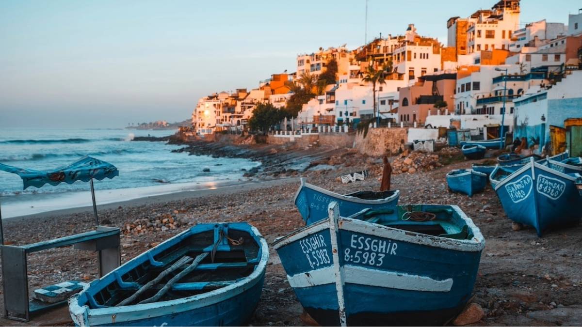 Blue fishing boats with white trim rest on a pebble beach in Taghazout, Morocco, as the sun sets over whitewashed houses climbing the hillside symbolizing the quiet rhythm of coastal life where wellness unfolds not through activity, but through presence and patience.