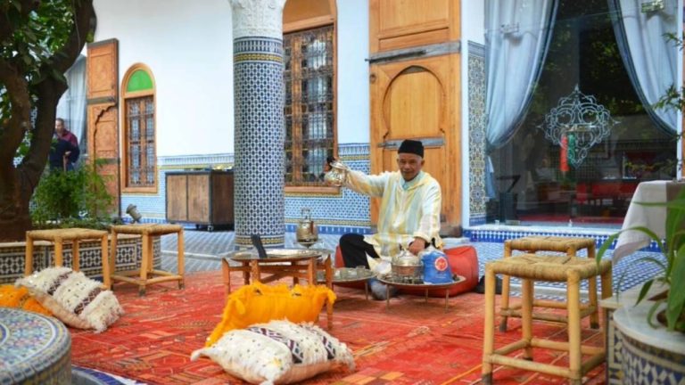 An Amazigh man in a striped djellaba pours mint tea from a silver pot into small glasses on a low table, seated on a red rug in a courtyard adorned with blue zellige tiles and wooden stools symbolizing the quiet rhythm of daily ritual where wellness unfolds not through performance, but through presence and patienc