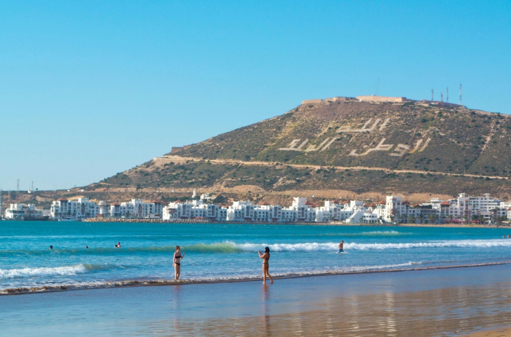 Turquoise waves lap at Agadir’s sandy shore as beachgoers enjoy the sun, while behind them, a hillside bears the name “أكادير”"Agadir" a reminder that beyond this postcard view lie ancient Amazigh traditions of argan, tea, storytelling, and celestial timekeeping that define the region’s true soul.