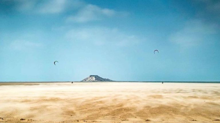 A lone kitesurfer glides across Dakhla’s endless sands and shallow waters under a vast blue sky, with a distant dune rising like an island symbolizing how the region’s traditions welcome strangers not as visitors, but as guests of the horizon itself.