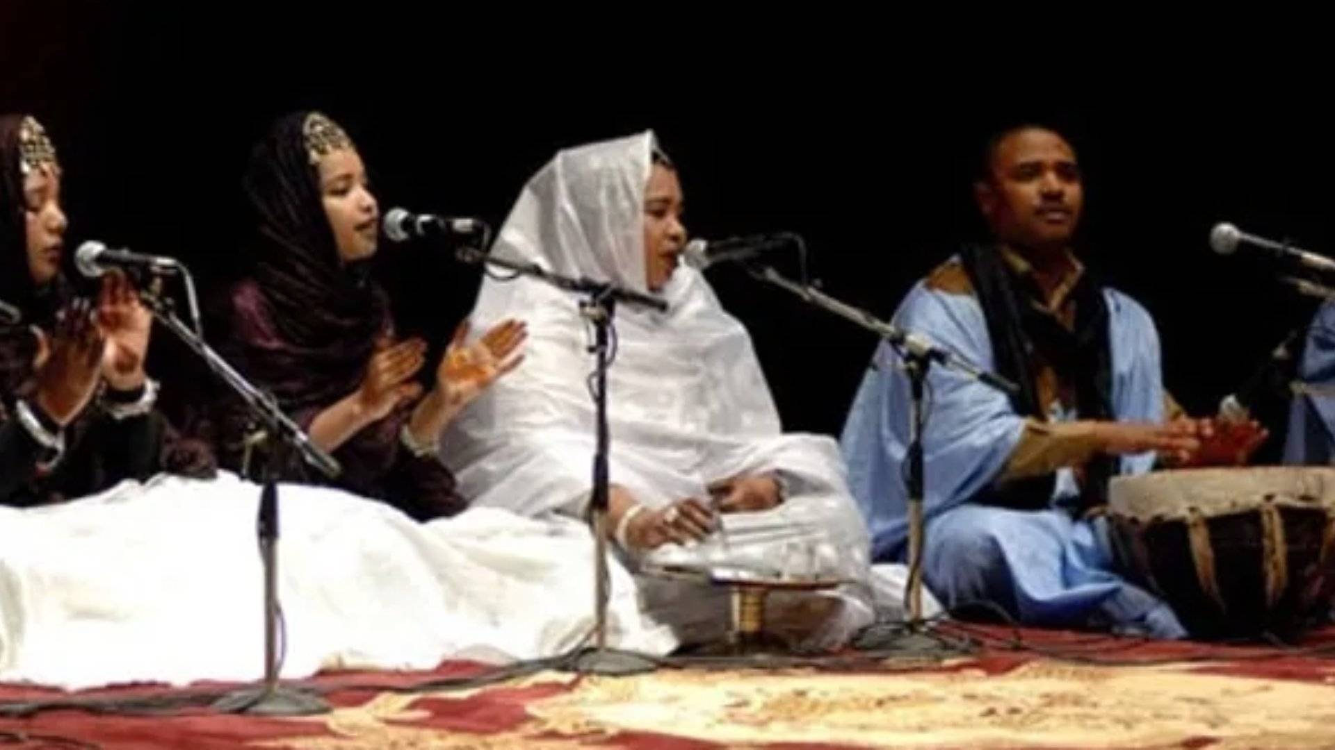 Three women in traditional Hassani dress sing madih chants into microphones while a man plays the tbal drum on stage in Dakhla symbolizing how sound, not spectacle, carries healing in a culture where rhythm follows the tide and community holds the song.