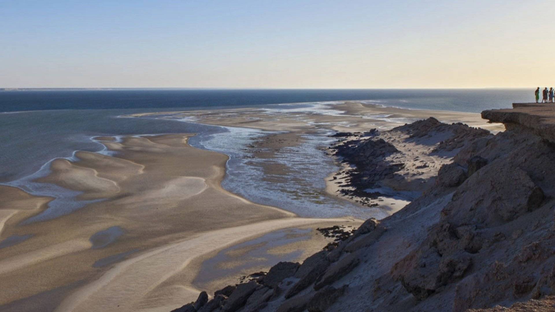 A lone figure stands on a rocky cliff overlooking Dakhla’s vast, empty beach at dawn, where sand and sea merge under a clear sky symbolizing how the region’s traditions measure time not in hours, but in tide, wind, and the quiet rhythm of presence.