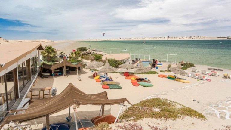 A beachfront lounge in Dakhla with colorful beanbags and thatched umbrellas overlooking turquoise waters and kitesurfers symbolizing how modern comfort coexists with ancient rhythm, where wellness flows not from luxury, but from presence aligned with wind, sand, and sea.