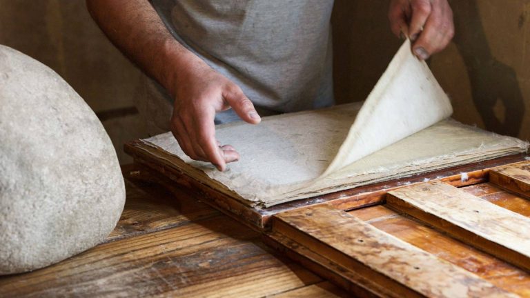 A craftsman’s hands lift a freshly formed sheet of handmade paper from a wooden frame in a traditional Fes workshop, with a stone weight and aged wooden tools nearby symbolizing how patience, texture, and touch form a living tradition where paper isn’t made, but allowed to become.