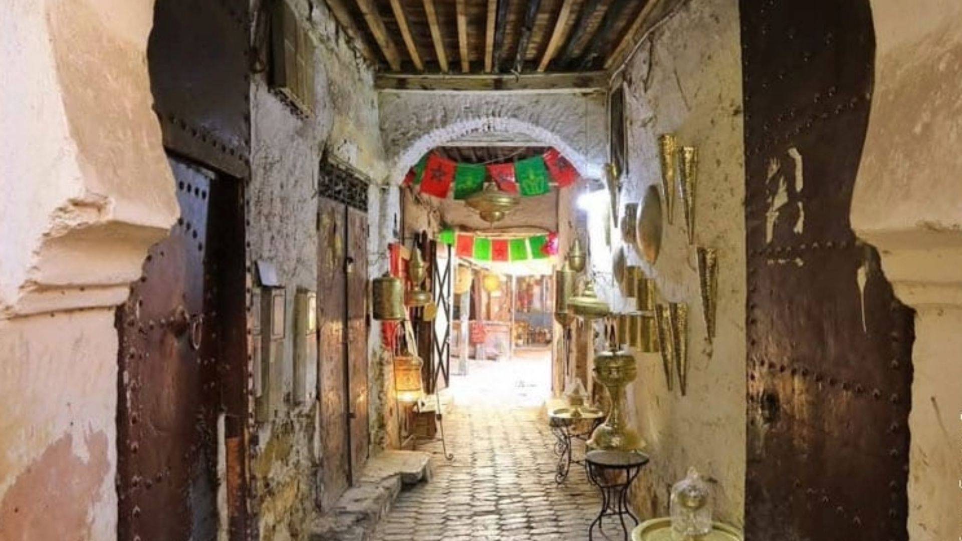 A sun-drenched alley in Fes’ medina, flanked by aged walls, iron-studded doors, and brass lanterns, with Moroccan flags strung overhead symbolizing how the city’s hidden passages don’t confuse the lost, but reveal them through scent, light, and the quiet rhythm of daily life.
