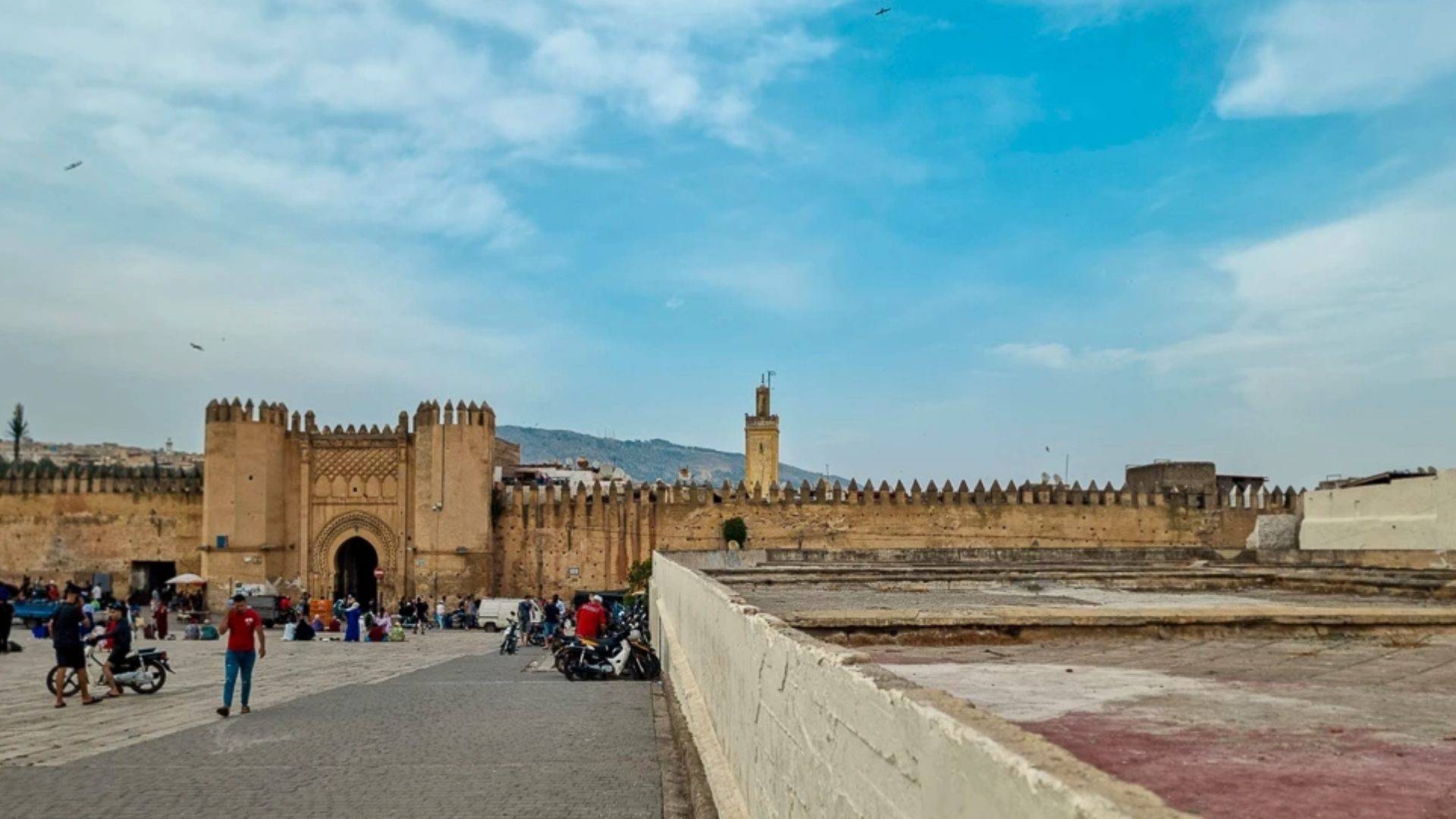 The historic Bab Jdid gate in Fes’ medina, with its fortified earth walls, crenellated ramparts, and distant minaret under a vast blue sky no tourist signs, no GPS markers, just people walking slowly, motorcycles parked, and the quiet invitation to step inside and lose yourself on purpose.